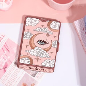 May include: A pink tarot card with a crescent moon, eye, and cloud design. The card reads "THE MOON" at the bottom and "XVIII" at the top. The card has a brown border and is surrounded by other pink items.