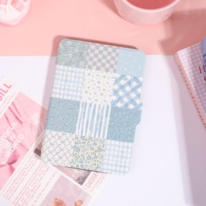 May include: A blue and white patchwork pattern notebook cover. The cover features various floral, gingham, and striped patterns in shades of blue and white. The notebook is open, revealing the interior pages. The words "BEAUTIFUL NEW WORLD" are visible on a magazine.
