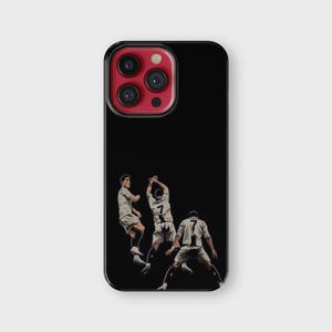 Puede incluir: Funda de tel&eacute;fono negra con una ilustraci&oacute;n estilizada de tres futbolistas en acci&oacute;n. Los jugadores visten uniformes blancos y negros. La funda tiene un borde negro y una lente de c&aacute;mara roja.
