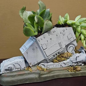May include: A white ceramic planter shaped like a Star Wars AT-ST walker with succulents growing out of the top. The planter is sitting on a brown base.