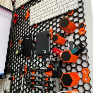 Może przedstawiać: Czarny organizer ścienny w kształcie plastra miodu z pomarańczowymi sześciokątnymi półkami. Organizer zawiera różne przedmioty, w tym dwie klawiatury, dysk twardy, zestaw długopisów, linijkę, słuchawki i różne inne narzędzia i akcesoria.