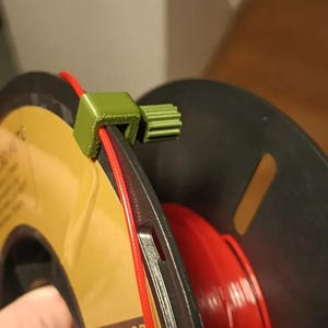 Può includere: Un clip guida per filamento stampato in 3D di colore verde attaccato a una bobina di filamento rosso. La bobina è etichettata "MAJKL3D filaments".