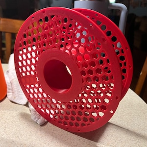 May include: A red plastic spool holder with a honeycomb pattern. The holder is designed to hold spools of filament for 3D printing.