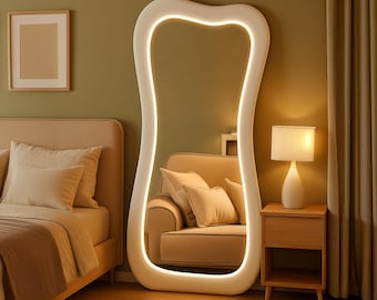 Puede incluir: Un espejo grande, blanco, de forma orgánica con iluminación integrada. El espejo está junto a una mesita de noche de madera con una lámpara. También se ve una cama y un sillón beige.
