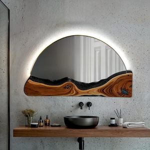 Può includere: Uno specchio da bagno moderno con una cornice in legno unica e una luce LED semicircolare. Lo specchio è montato su una parete in cemento sopra un lavabo nero.
