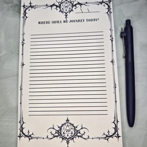Puede incluir: Un bloc de notas rectangular de papel color hueso con bordes de tinta negra, una ilustración de un dado de veinte caras en la parte superior e inferior, y el texto WHERE SHALL WE JOUNREY TODAY? sobre líneas horizontales, junto a un bolígrafo azul.