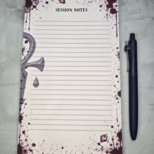 Puede incluir: Un bloc de notas rectangular con líneas horizontales y el texto SESSION NOTES en la parte superior. El borde presenta un símbolo ankh gris, salpicaduras de color rojo sangre e ilustraciones de dados. Un bolígrafo azul oscuro está al lado.