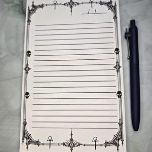 Puede incluir: Un bloc de notas rectangular blanco con líneas horizontales negras y un borde decorativo con símbolos ankh, filigranas ornamentadas y pequeñas calaveras. Un bolígrafo retráctil negro está colocado verticalmente a la derecha del bloc.