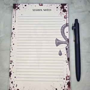 Puede incluir: Un bloc de notas rectangular titulado SESSION NOTES con líneas horizontales, que presenta un diseño de salpicaduras de sangre de color rojo oscuro y un símbolo ankh gris en el lado derecho, junto a un bolígrafo negro mate retráctil.