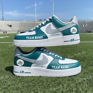 May include: Two custom teal, white, and gray sneakers with the text "TEAM NAME" and "CUSTOM NAME" on the sides. The shoes have a white sole with "AIR" printed on the side. The sneakers are on a green field.