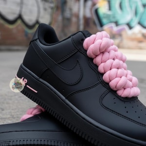 Może przedstawiać: Czarne buty sportowe Air Force 1 z r&oacute;żowymi plecionymi sznurowadłami. Buty mają czarną podeszwę, logo Nike i napis "AIR" w kolorze r&oacute;żowym z boku. Tło to rozmyta scena miejska.