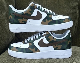 May include: A pair of white-soled sneakers with a camouflage pattern in shades of green, brown, and tan. The shoes feature a brown Nike swoosh and white laces. The word "AIR" is visible on the midsole.