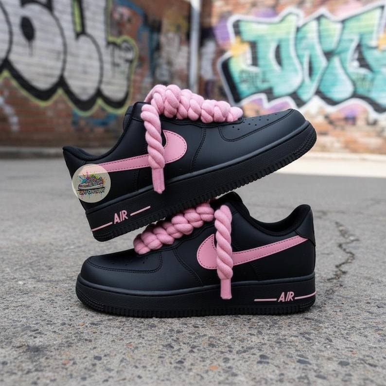 Może przedstawiać: Czarne sneakersy z r&oacute;żowymi akcentami, w tym logo Nike i sznurowadłami. Słowo "AIR" jest wydrukowane na r&oacute;żowo z boku. Buty są ułożone jeden na drugim, na tle graffiti.