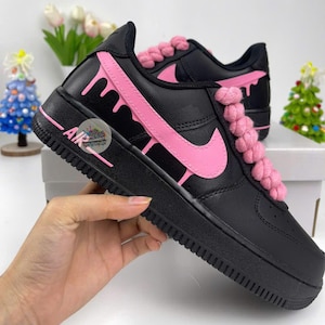 Może przedstawiać: Czarne sneakersy z r&oacute;żowymi akcentami, w tym wzorem spływającej farby i r&oacute;żowym logo Nike. Sznurowadła są grube i r&oacute;żowe. Słowo "AIR" jest wydrukowane na boku podeszwy.