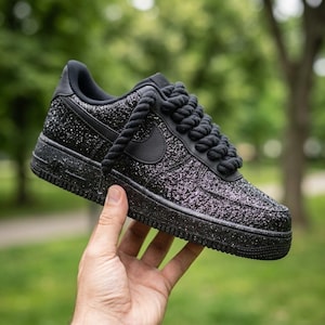 May include: Black Air Force 1 sneakers with a glittery finish and thick rope laces. The shoe features a black Nike swoosh and a textured sole. The shoe is held up against a blurred green background.