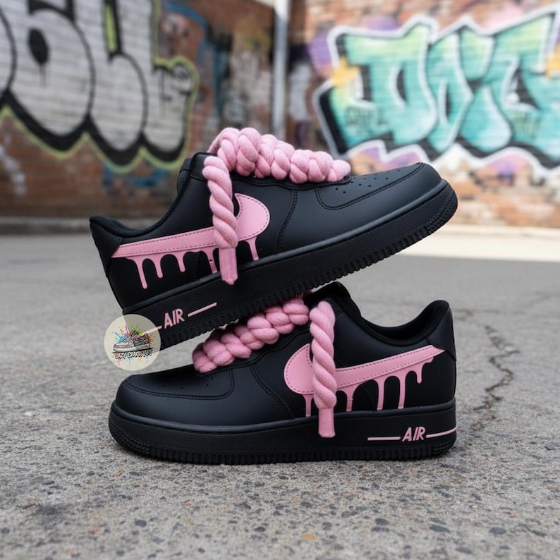 May include: Black sneakers with pink accents, including a dripping design on the side and a pink swoosh. The laces are replaced with thick, pink rope. The word "AIR" is printed on the side of the sole. The shoes are stacked on top of each other.
