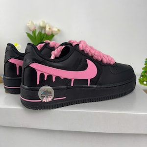 Może przedstawiać: Czarne i r&oacute;żowe sneakersy Air Force 1 z r&oacute;żowym logo Nike i r&oacute;żowym wzorem spływającej farby. Sneakersy mają r&oacute;żowe sznurowadła i czarną podeszwę. Z boku nadrukowano napis "AIR".