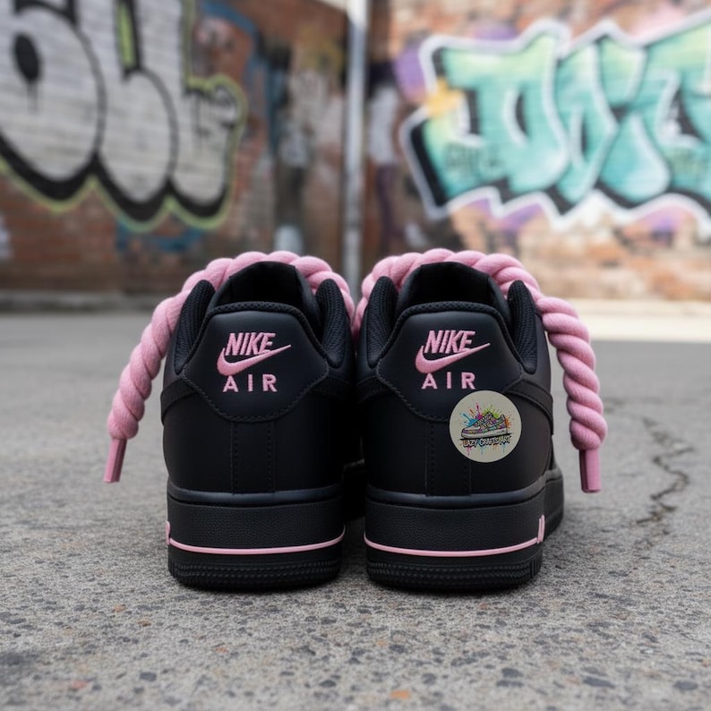 Może przedstawiać: Para czarnych sneakers&oacute;w z r&oacute;żowymi akcentami, w tym sznurowadłami i logo Nike Air. Buty mają niski kr&oacute;j i naklejkę z boku. Tło przedstawia graffiti na ceglanej ścianie.