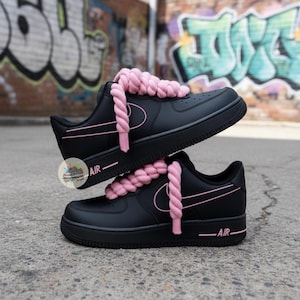 Może przedstawiać: Czarne sneakersy z r&oacute;żowymi akcentami i grubymi r&oacute;żowymi sznurowadłami. Buty mają klasyczny design z napisem "AIR" z boku. Tło przedstawia graffiti.
