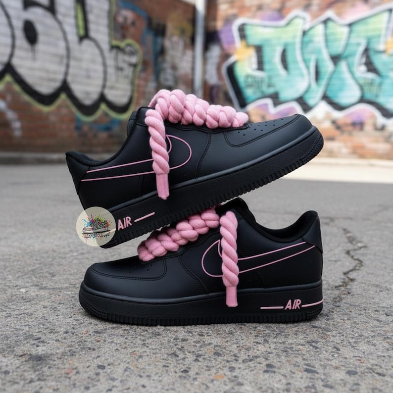 Może przedstawiać: Czarne sneakersy z r&oacute;żowymi akcentami i grubymi r&oacute;żowymi sznurowadłami. Buty mają klasyczny design z napisem "AIR" z boku. Tło przedstawia graffiti.