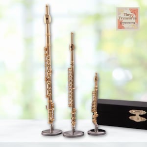 May include: Three miniature gold-coloured flutes on stands, with a black wooden box in the background. The flutes are different sizes, with the largest flute being the tallest. The text "Tiny Treasures" is on the box.