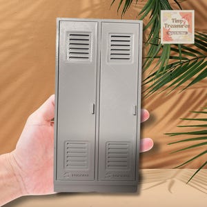 May include: A grey miniature locker with two doors, each featuring a vent and a handle. The bottom of the locker has the text "ARSENAL". The locker is held in a hand.