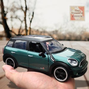 Könnte beinhalten: Grünes Spielzeugautomodell eines Mini Cooper Countryman. Das Auto hat schwarze Räder und ein schwarzes Dach. Das Auto steht auf der Hand einer Person.