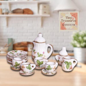 Puede incluir: Un juego de té de porcelana en miniatura con base blanca y estampado floral. El juego incluye una tetera, una jarra de crema, un azucarero y cuatro tazas de té y platillos. El juego se muestra sobre una superficie blanca.