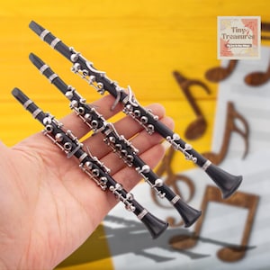 Op de afbeelding: Drie miniatuur zwarte klarinetten met zilveren toetsen worden in een hand tegen een gele achtergrond gehouden. De klarinetten zijn allemaal even groot en hebben een vergelijkbaar ontwerp. De achtergrond heeft muzieknoten.