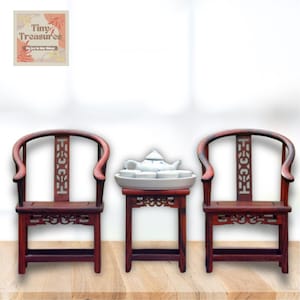 May include: A set of three miniature wooden furniture pieces, including two chairs and a table. The chairs are dark red with intricate carvings and curved backs. The table is also dark red with a square top and four legs. A small white teapot and cups are on the table.