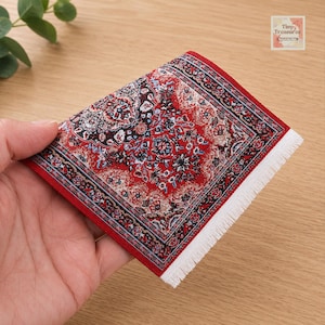 Puede incluir: Una alfombra rectangular en miniatura con un detallado patrón floral en rojo, negro y blanco, con un medallón central y un borde intrincado. El tejido tiene flecos blancos en un lado. El texto Tiny Treasures es visible.