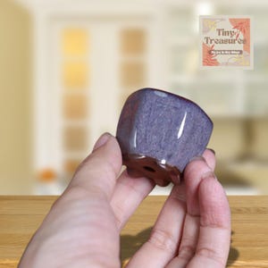 May include: A small, purple ceramic planter with a square shape and a drainage hole. The planter is glazed with a light purple glaze and has a brown rim.