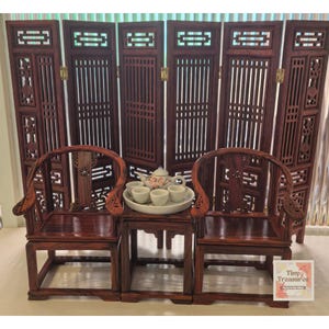 May include: A miniature wooden tea set with two chairs and a table, set in front of a dark wood room divider. The tea set includes a teapot and four cups.