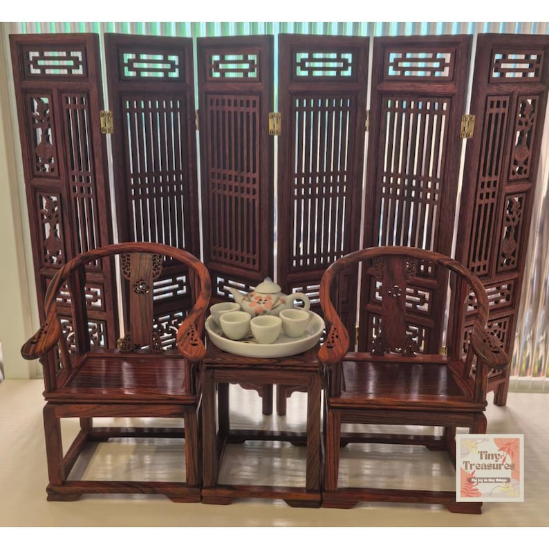 May include: A miniature wooden tea set with two chairs and a table, set in front of a dark wood room divider. The tea set includes a teapot and four cups.