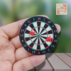 May include: A small dartboard with red darts. The dartboard has black and white segments with red and green details. Numbers 1 to 20 are around the edge. The image includes the text "Tiny Treasures".