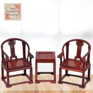 May include: A set of three miniature wooden furniture pieces, including two chairs and a table. The chairs have a curved back and intricate carvings. The table is square and has a simple design. The furniture is made of a dark reddish-brown wood.