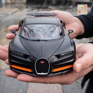 Può includere: Un'auto giocattolo nera opaca con dettagli arancioni, tenuta in mani aperte. L'auto presenta uno spoiler, fari dettagliati e il logo Bugatti. Il design è elegante e aerodinamico, una replica in miniatura di un veicolo di lusso.