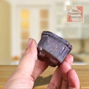 Puede incluir: Una pequeña maceta cuadrada de cerámica con un esmalte púrpura brillante. La maceta tiene un borde ligeramente ensanchado y pequeños orificios de drenaje. La imagen incluye el texto "Tiny Treasures" y "Big joy in tiny things".