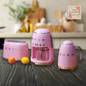 Op de afbeelding: Een set van drie roze miniatuur keukenapparaten: een broodrooster, een koffiezetapparaat en een waterkoker. Elk apparaat heeft het woord "SMEG" in zwarte letters op de voorkant.