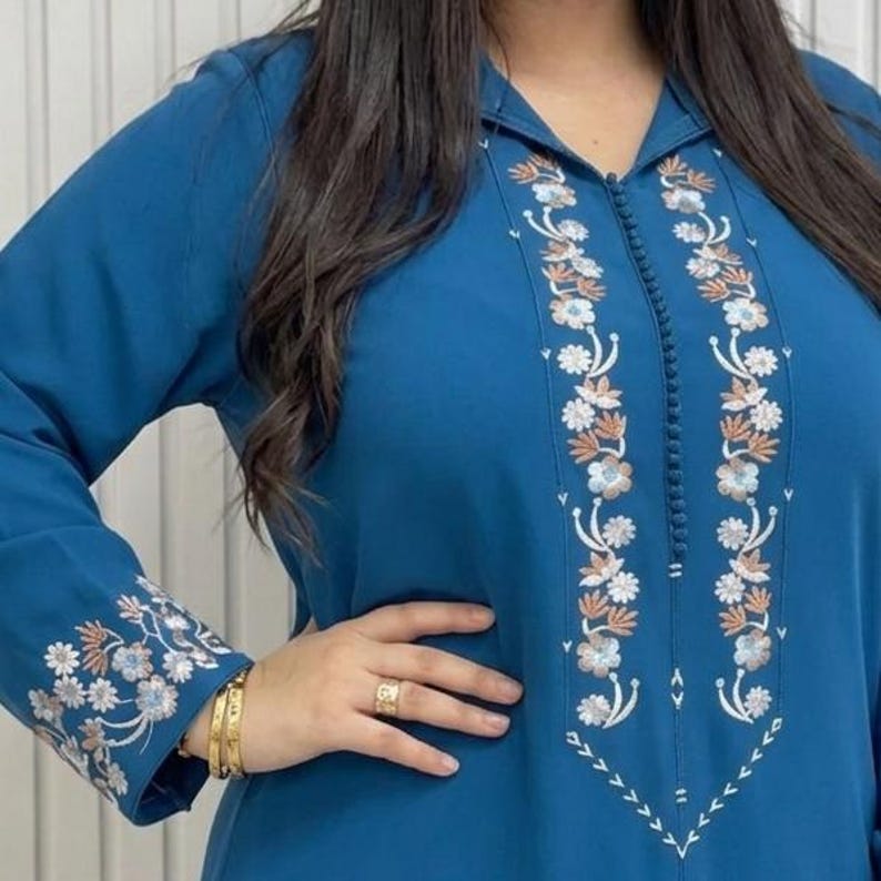 May include: A teal blue tunic with long sleeves and floral embroidery. The tunic features white, orange, and light blue floral designs along the front and sleeve cuffs. The collar is a classic V-neck style.