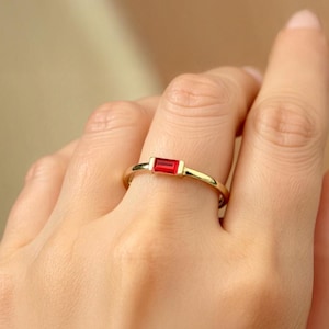 May include: A gold ring with a rectangular, bright red gemstone. The ring is simple and elegant, with a thin band. The gemstone is set horizontally in the center of the ring.