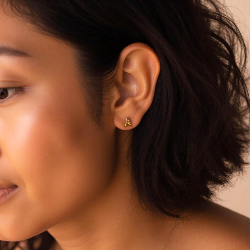 K&ouml;nnte beinhalten: Nahaufnahme eines Ohrs mit einem goldenen Initial 'A' Ohrstecker.