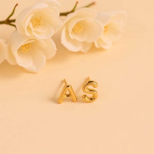 K&ouml;nnte beinhalten: Ein Paar goldene Initialohrringe, einer mit dem Buchstaben "A" und der andere mit dem Buchstaben "S".