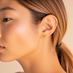 K&ouml;nnte beinhalten: Nahaufnahme eines Ohrs mit einem goldenen Initial-Ohrring 'M'. Der Ohrring ist schlicht und minimalistisch gestaltet.