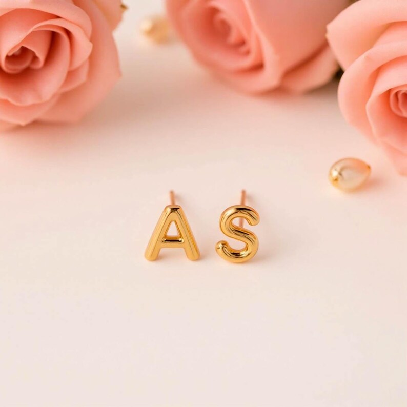 K&ouml;nnte beinhalten: Ein Paar goldene Initialohrringe mit den Buchstaben A und S. Die Ohrringe befinden sich auf einem wei&szlig;en Hintergrund mit rosa Rosen.