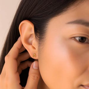 K&ouml;nnte beinhalten: Nahaufnahme eines Ohrs mit einem goldenen Initial 'A' Ohrstecker.