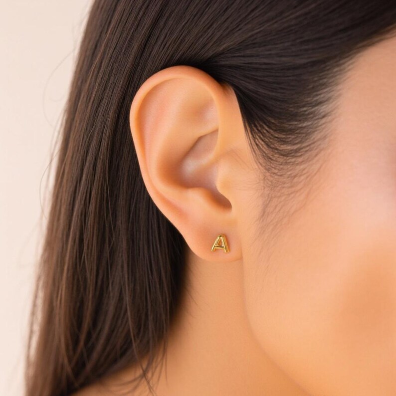 K&ouml;nnte beinhalten: Nahaufnahme eines Ohrs mit einem goldenen Initial 'A' Ohrstecker.