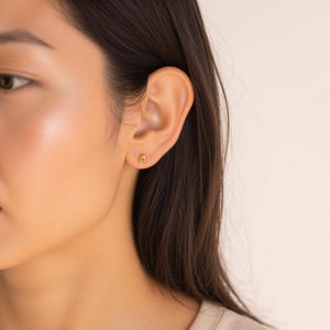 Może przedstawiać: Zbliżenie na ucho osoby z małym złotym kolczykiem w kształcie litery 'A'.