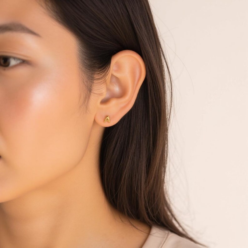 K&ouml;nnte beinhalten: Nahaufnahme eines Ohrs mit einem kleinen goldenen Buchstaben 'A' als Ohrstecker.