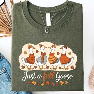 a green t-shirt with a graphic design of four geese wearing orange sweaters and hats, along with the text "Just a fall goose". The t-shirt is displayed on a white background, accompanied by a pair of blue jeans, a pair of white sneakers, and some dried leaves.
